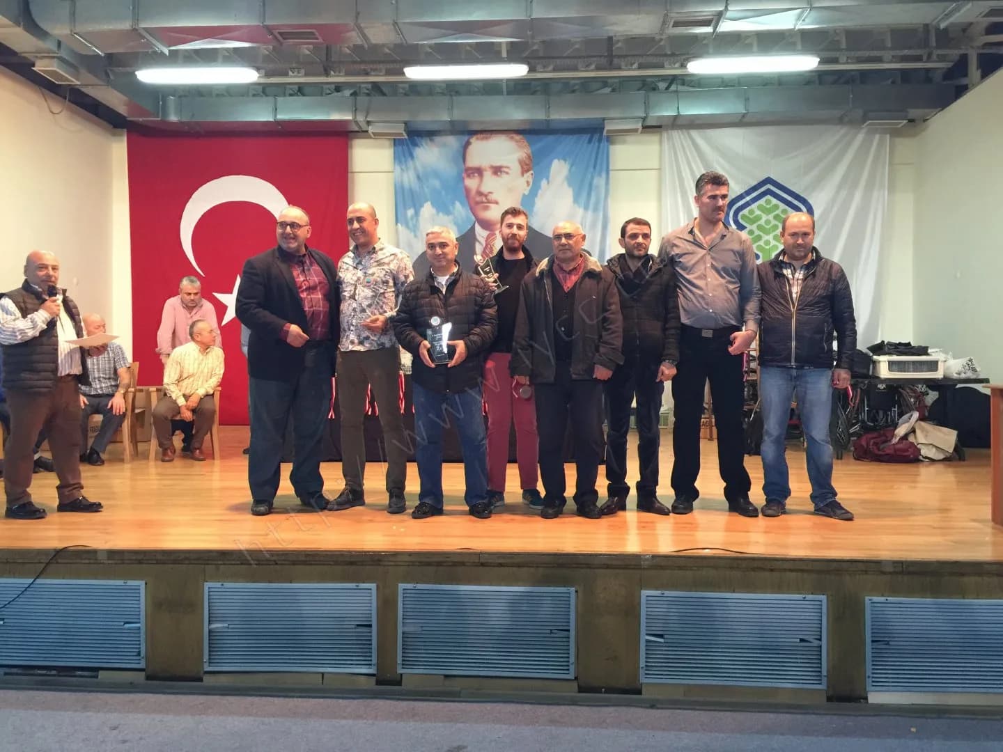 IYCC 2017 ödül töreninde soldaki kişi mikrofonla anons yaparken sahnede plaketleriyle sıralanan grup