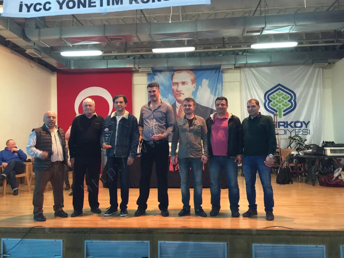 IYCC 2017 ödül töreninde sahnede sekiz kişi, ikisi cam plaket tutuyor, arkada Türk bayrağı ve Atatürk posteri