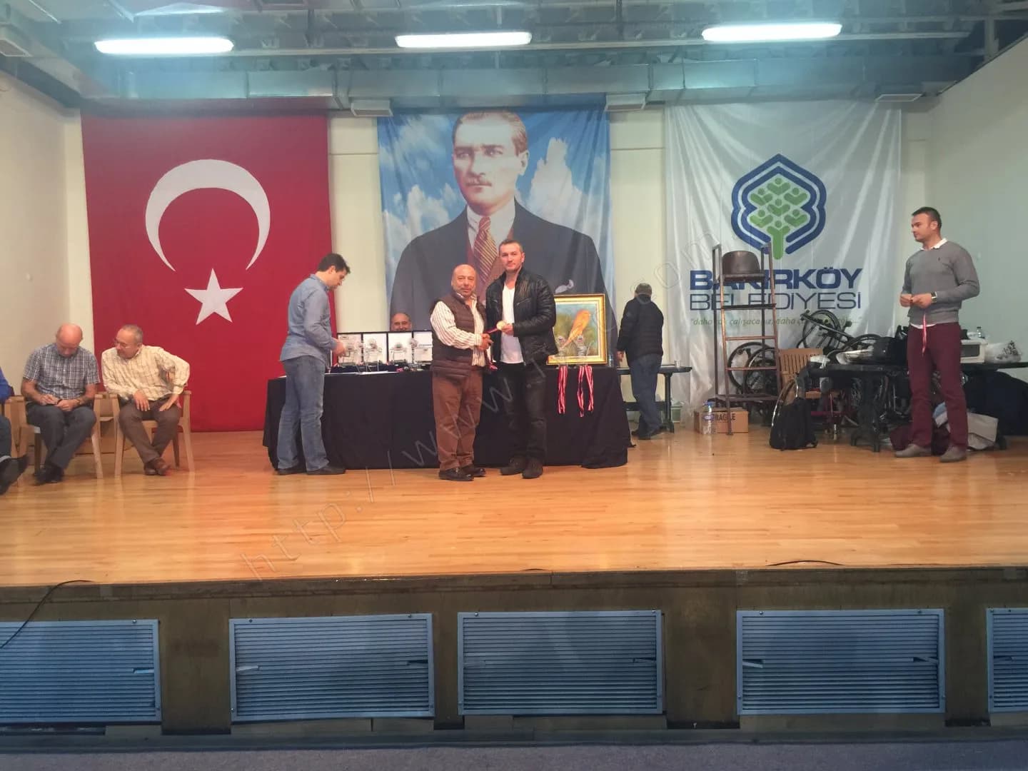 IYCC 2017 ödül töreninde sahnenin ortasında madalya takdimi, masada plaketler ve çerçeveli kanarya tablosu