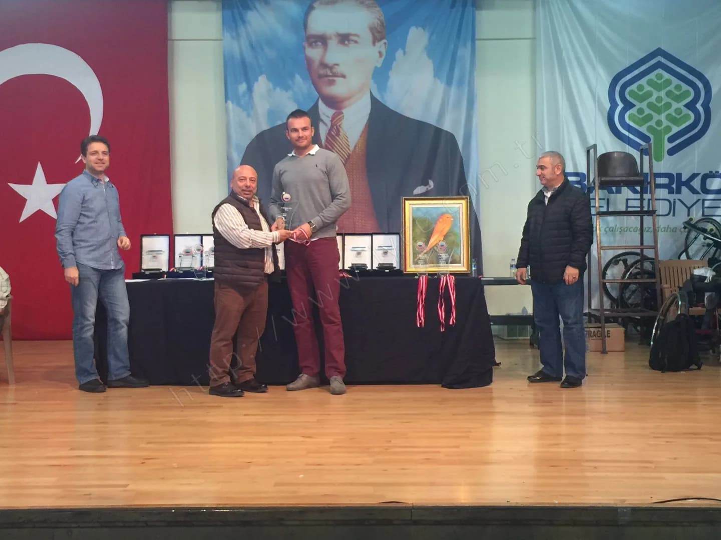 IYCC 2017 ödül töreninde plaket takdimi; masada turuncu bir kanaryanın resmedildiği çerçeveli tablo duruyor