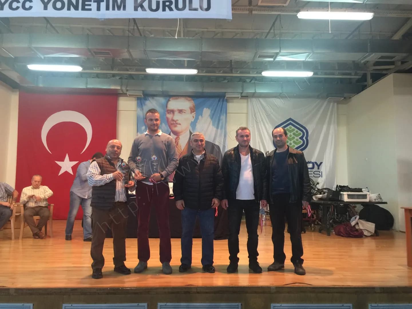 IYCC 2017 ödül töreninde sahnede beş kişi, gri kazaklı katılımcı iki cam plaketi birlikte tutuyor