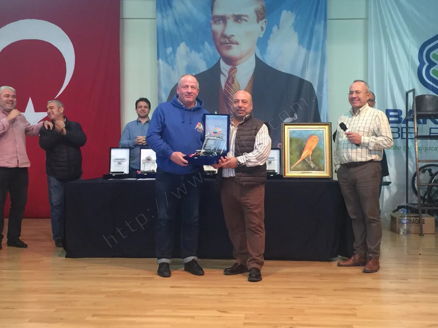 IYCC 2017 ödül töreninde kutulu plaket takdimi yakından; yanda mikrofonlu sunucu ve kanarya tablosu