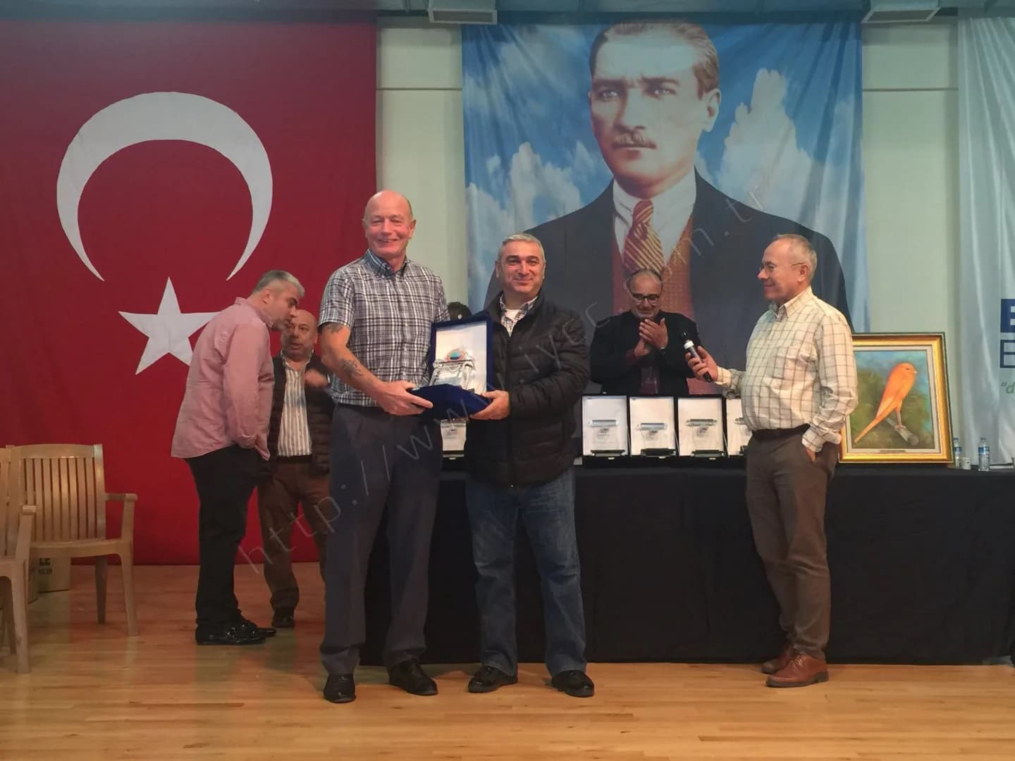 IYCC 2017 ödül töreninde ekose gömlekli katılımcı kutulu plaketini alıyor, sunucu mikrofonla konuşuyor