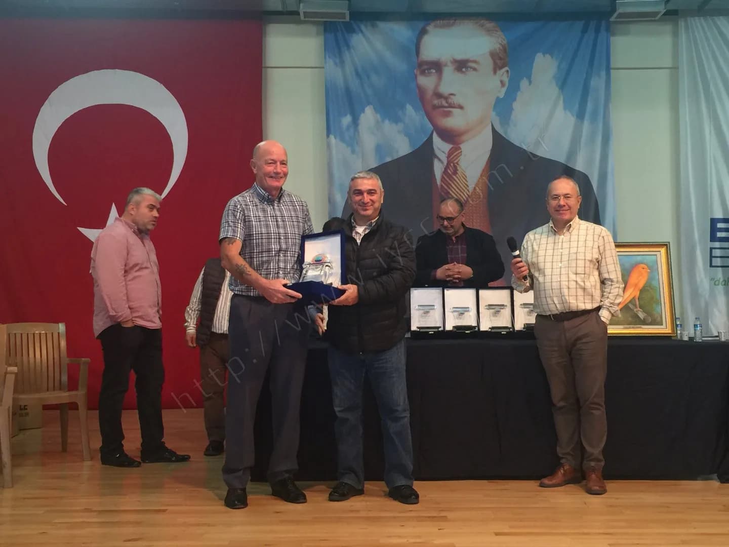 IYCC 2017 ödül töreninde iki kişi kutulu plaketi birlikte tutarak objektife gülümsüyor, arkada kanarya tablosu