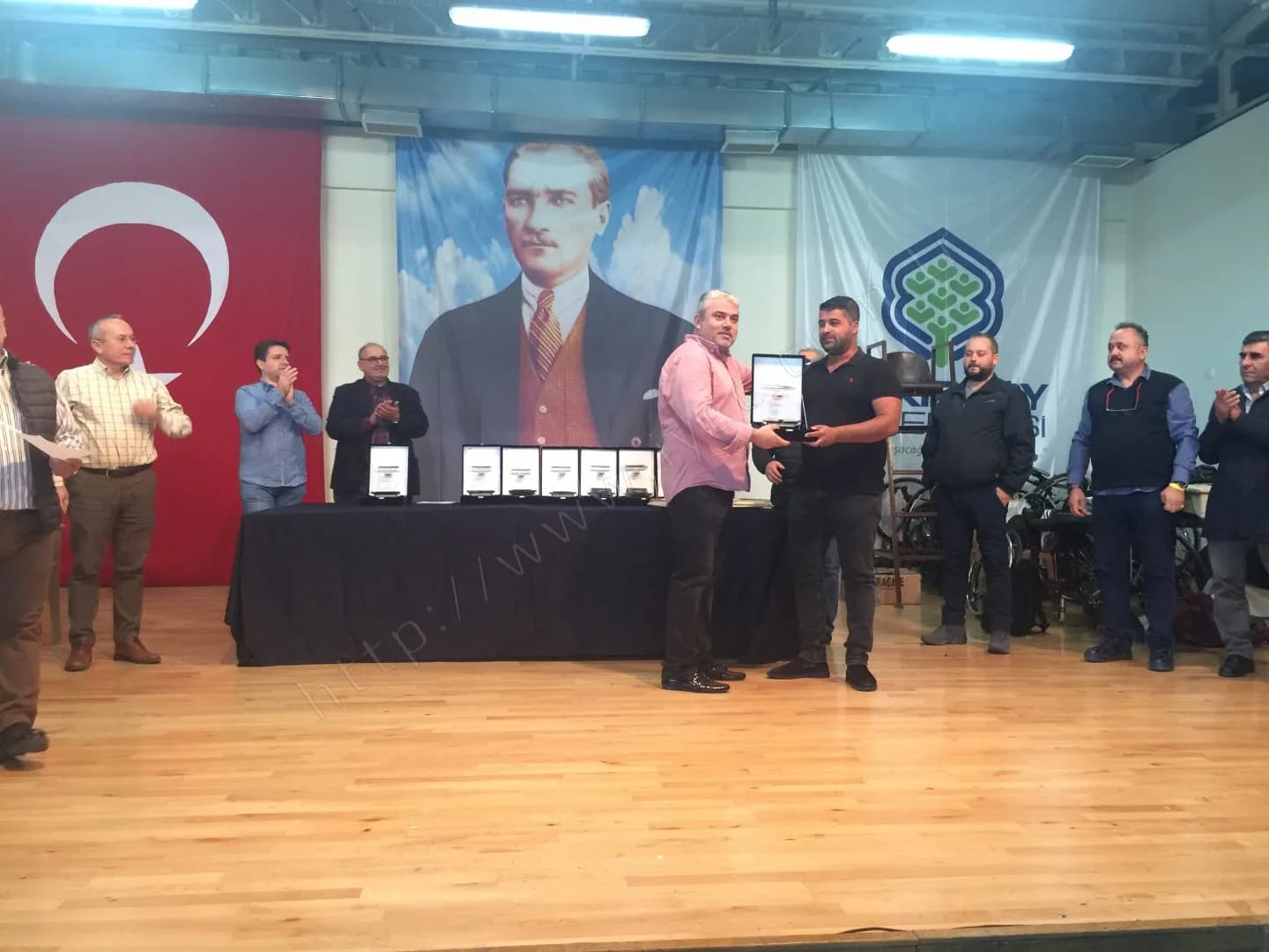 IYCC 2017 ödül töreninde sahnede plaket teslim ediliyor, iki yandakiler alkışlıyor, masada sıralı plaketler