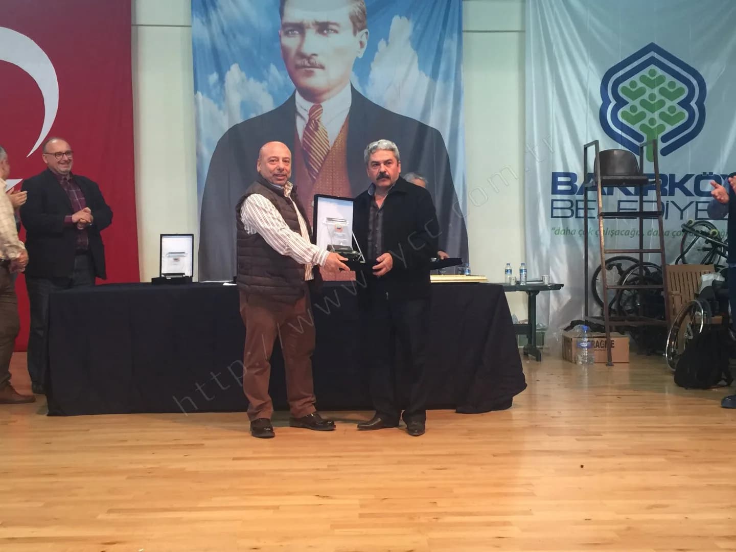 IYCC 2017 ödül töreninde yelekli görevli bıyıklı katılımcıya plaket veriyor, solda alkışlayanlar var