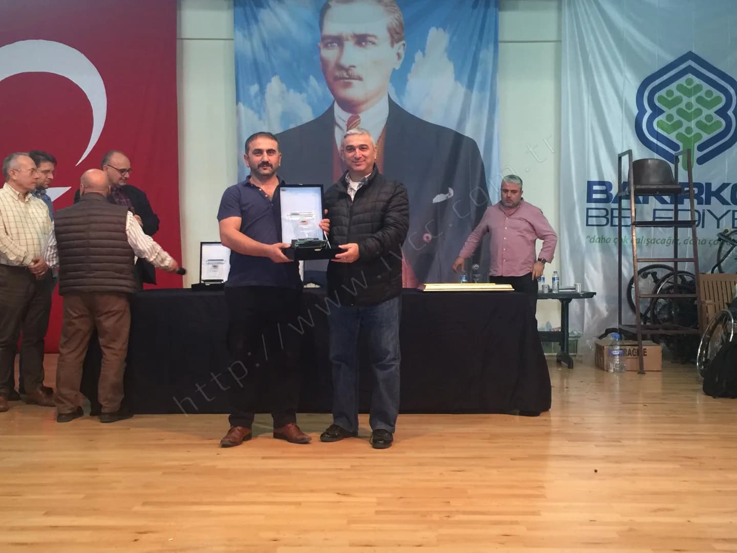 IYCC 2017 ödül töreninde lacivert tişörtlü katılımcı ile şişme montlu görevli plaketi birlikte tutuyor