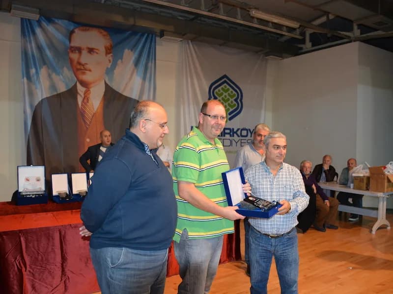 A man in a green striped shirt hands a plaque in a blue box to another man at the IYCC 2018 final, spectators behind