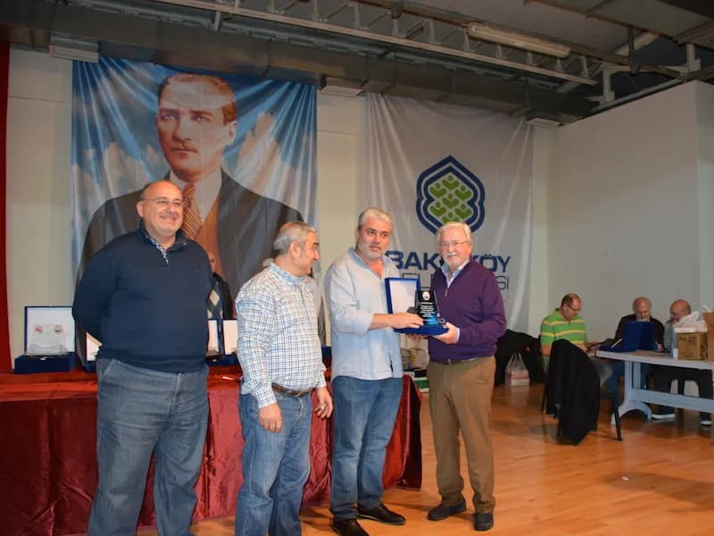 A man in a purple sweater is handed a plaque in a blue box at the IYCC 2018 final, with seated spectators behind