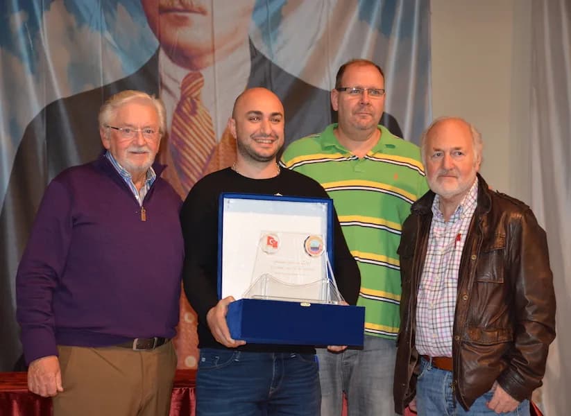 Four men pose at the IYCC 2018 final; the one in the centre holds a large crystal trophy in a blue box with both hands