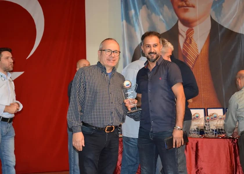 Two men smile holding a crystal trophy together at the IYCC 2018 awards, with the Turkish flag and award table behind