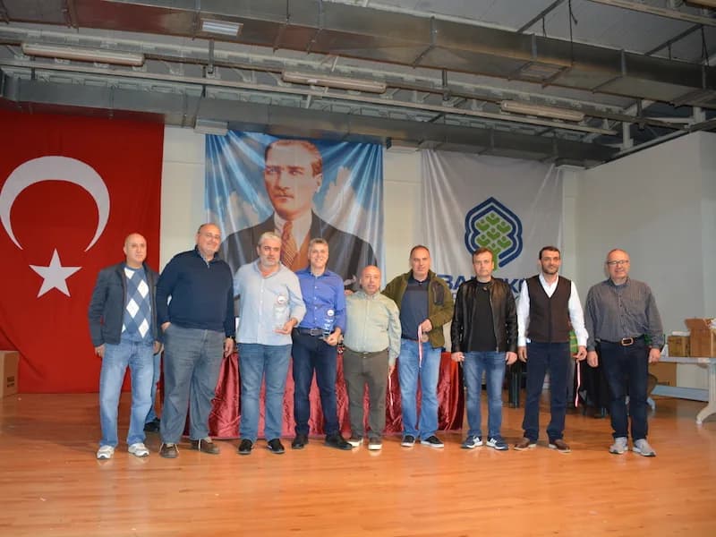 Nine men pose side by side before the Turkish flag and an Atatürk portrait at the IYCC 2018 final, one holding a trophy