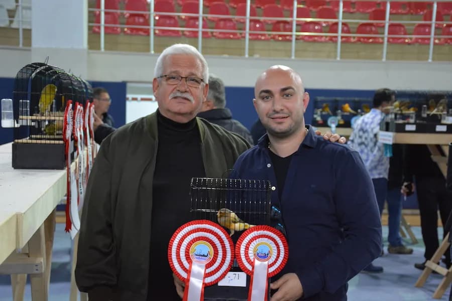 IYCC 2018 yarışmasında iki kişi, iki kırmızı-beyaz ödül rozeti takılı ve içinde sarı kanarya olan kafesle poz veriyor